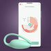 The Elvie Trainer Smart Kegel Exerciser in mint green sits in front of a smartphone displaying workout stats and pelvic floor strength summaries on its app, all set against a purple background.