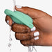 A hand with white-painted nails rinses the green, oval-shaped Elvie Trainer Smart Kegel Exerciser under running water against a plain white background, showcasing this essential tool for pelvic floor strength.