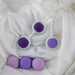 Four round, purple and lavender weighted kegel balls with two white holders featuring circular slots rest on soft, white fabric—ideal for pelvic floor strengthening with the Wellness Kegel Ball Training Kit for Pelvic Floor Health.