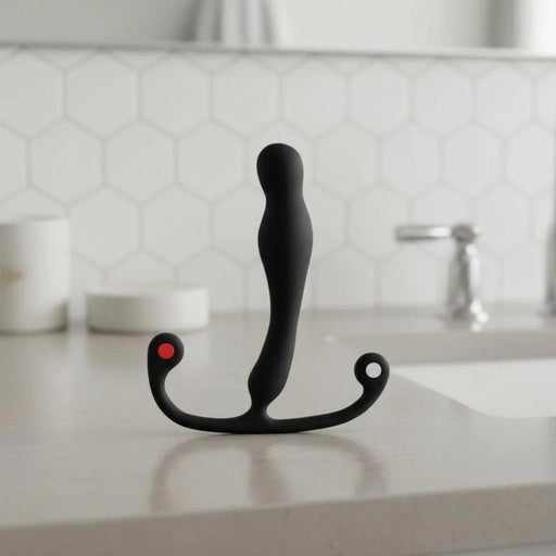 The Aneros Eupho Syn Trident Hands-Free Silicone Prostate Massager, black and curved with two ring-shaped handles and a red dot, stands upright on a bathroom countertop with hexagonal tile backsplash and a faucet in the background.
