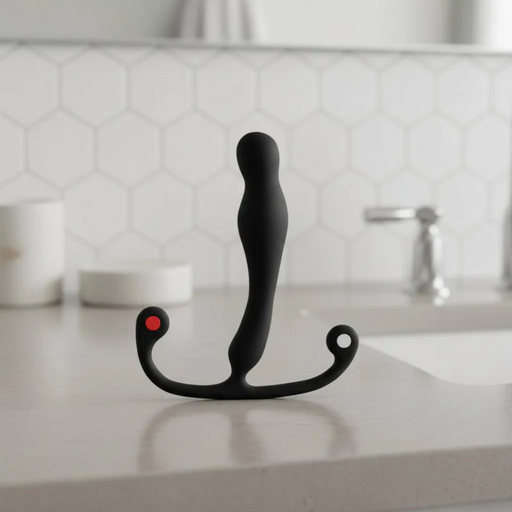 The Aneros Eupho Syn Trident Hands-Free Silicone Prostate Massager, black and curved with two ring-shaped handles and a red dot, stands upright on a bathroom countertop with hexagonal tile backsplash and a faucet in the background.