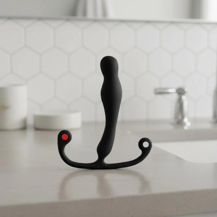 The Aneros Eupho Syn Trident Hands-Free Silicone Prostate Massager, black and curved with two ring-shaped handles and a red dot, stands upright on a bathroom countertop with hexagonal tile backsplash and a faucet in the background.