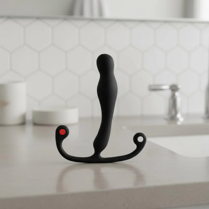 The Aneros Eupho Syn Trident Hands-Free Silicone Prostate Massager, black and curved with two ring-shaped handles and a red dot, stands upright on a bathroom countertop with hexagonal tile backsplash and a faucet in the background.