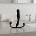 The Aneros Eupho Syn Trident Hands-Free Silicone Prostate Massager, black and curved with two ring-shaped handles and a red dot, stands upright on a bathroom countertop with hexagonal tile backsplash and a faucet in the background.