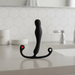 The Aneros Eupho Syn Trident Hands-Free Silicone Prostate Massager, black and curved with two ring-shaped handles and a red dot, stands upright on a bathroom countertop with hexagonal tile backsplash and a faucet in the background.