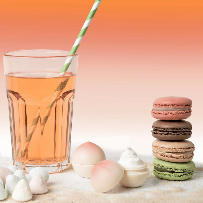 A swirl of cream sits beside pastel macarons, dome-shaped candies, and a peach drink with a green-and-white striped straw—all on a light surface with an orange gradient background, featuring Exsens Flavored Nipple Arousal Cream - Peachy Keen (8ml), vegan and paraben free.