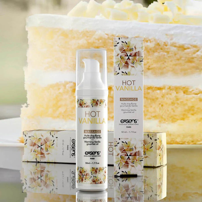 A bottle and box of Exsens Hot Vanilla Flavored Warming Massage Oil 50ml, featuring white and floral packaging with gold text, are displayed in front of a layered vanilla cake with white frosting, hinting at this oral pleasure enhancer.