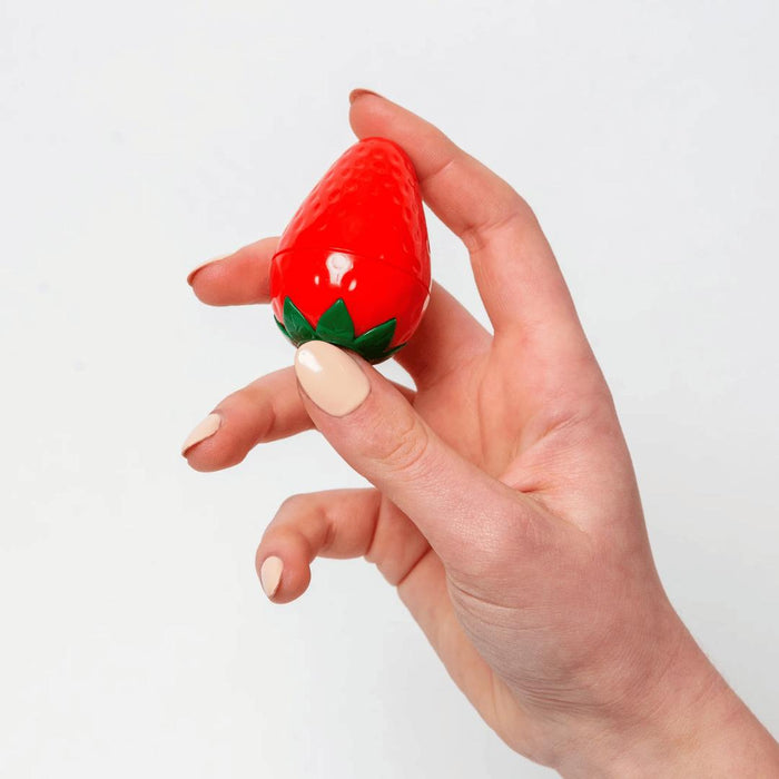 A hand with light skin holds the Exsens Oh My Strawberry Flavored Nipple Arousal Cream (8ml), shaped like a small red strawberry, against a white background. This vegan formula is also paraben free.