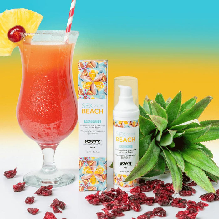 A tropical cocktail garnished with pineapple and cherry sits beside a bottle of Exsens Sex on the Beach Flavored Warming Massage Oil 50ml, surrounded by pineapple top and dried cranberries, on a yellow-blue gradient background.