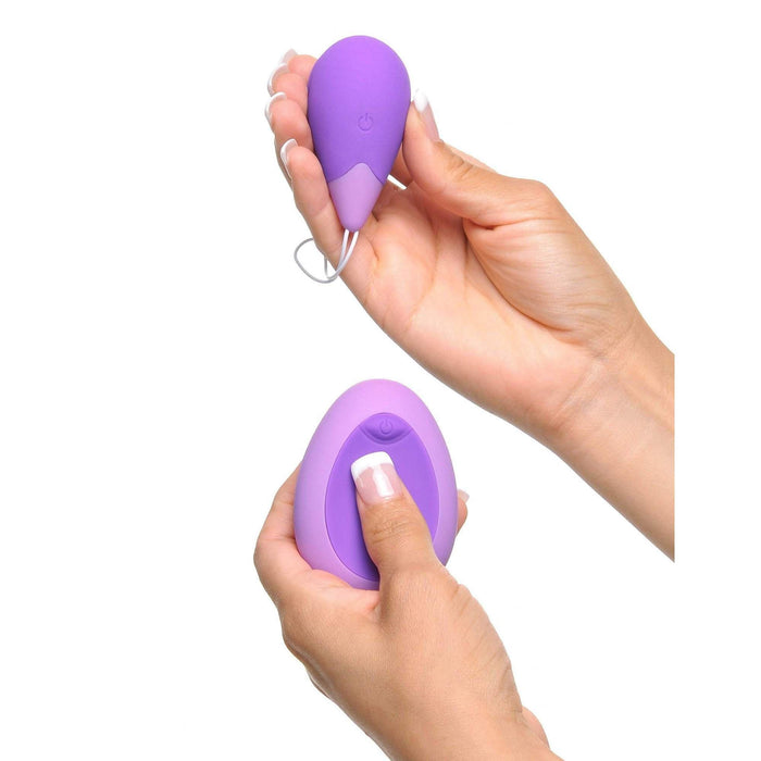 A person holds the compact, purple Fantasy For Her Remote Controlled Kegel Exerciser from Pipedream Products in one hand and its matching remote control with an attached white cord in the other hand, all set against a plain white background.