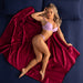 A woman in lilac lingerie lies on a navy bed covered by the Liberator Fascinator Throw Queen Size Waterproof Sex Blanket, striking a relaxed, confident pose with one arm behind her head and her eyes closed.