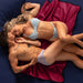 A couple in underwear lies closely together on a Liberator Fascinator Travel Sized Waterproof Sex Blanket, smiling and touching affectionately. The blanket’s red color stands out against the dark blue bedding background.
