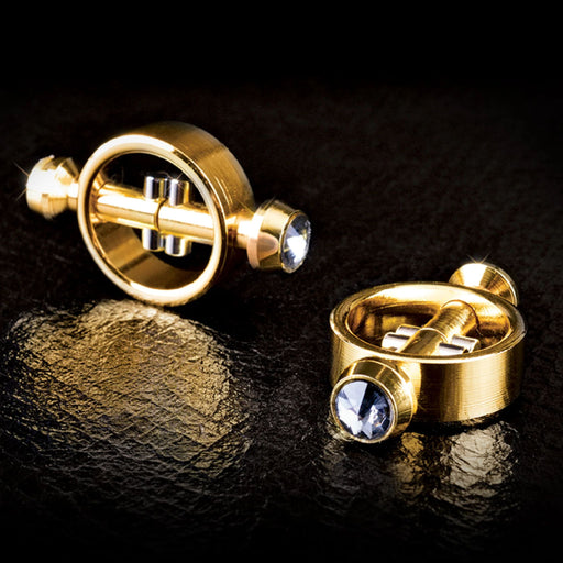 A pair of Fetish Fantasy Gold Magnetic Nipple Clamps with Diamond Tips from Pipedream Products placed on a dark surface. The clamps feature an elegant design and are adorned with faux diamond tips, giving them a luxurious and sophisticated appearance.