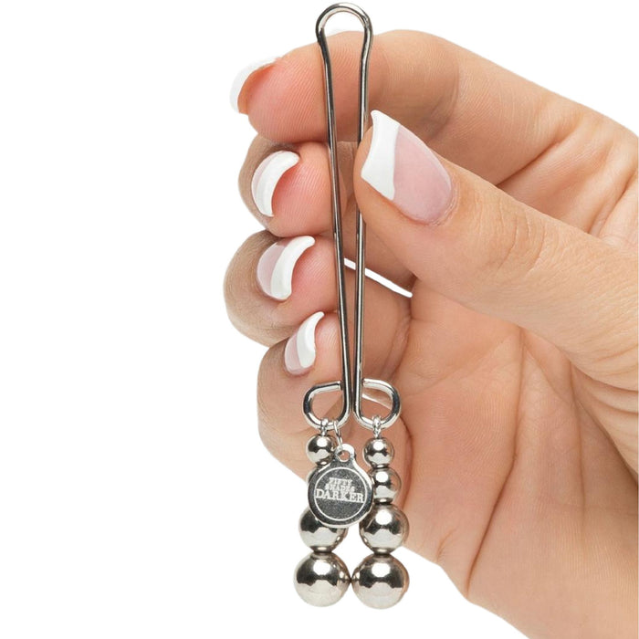 A hand with manicured nails holding a Fifty Shades of Grey Just Sensation Beaded Clitoral Clamp, featuring three attached metal beads and reminiscent of refined elegance. A logo featuring the word “DALLE” is imprinted on a circular part near the beads. The background is white.
