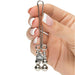 A hand with manicured nails holding a Fifty Shades of Grey Just Sensation Beaded Clitoral Clamp, featuring three attached metal beads and reminiscent of refined elegance. A logo featuring the word “DALLE” is imprinted on a circular part near the beads. The background is white.