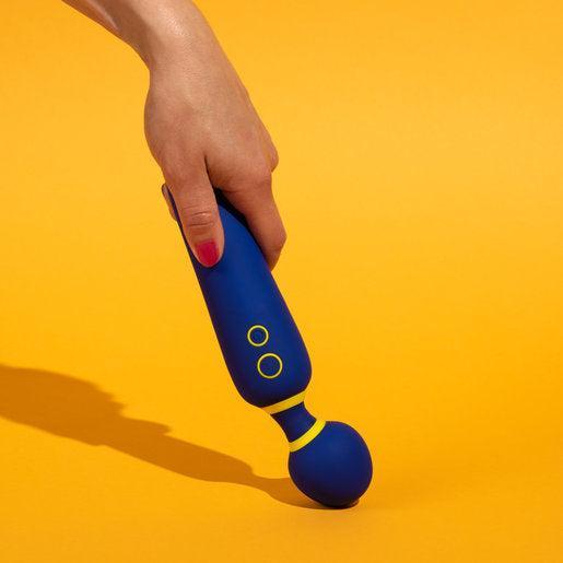 A hand with pink nail polish is holding the Flip Powerful Beginner's Wand Vibrator by WOW, a blue, USB rechargeable vibrator featuring a rounded top and two yellow buttons on its body, all set against a bright orange background.