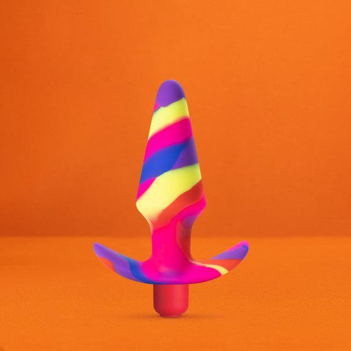 A striking, cone-shaped silicone object with a flared base, showcasing a lively swirl of pink, blue, yellow, and purple hues, is displayed against an orange background. Named the "Free Spirit Vibrating 10 Function Beginner Silicone Butt Plug" by Blush, this sex toy is made from platinum-cured silicone and stands upright, making it perfect for beginners.