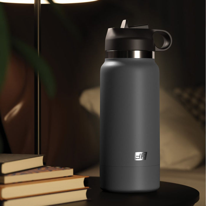 A black matte insulated **Fuck Flask Pussy Stroker Disguised as a Water Bottle - Vanilla** by **Pipedream Products** with a lid and handle sits discreetly on a black table next to a small stack of books. A nearby lamp casts a warm light over the scene, creating a cozy ambiance. In the background, a pillow and part of a sofa are visible.