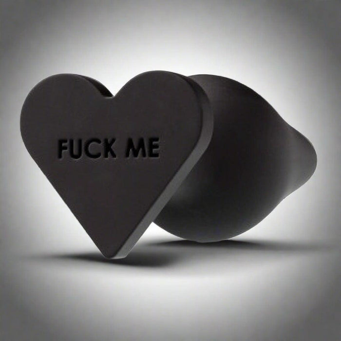 A black, heart-shaped Fuck Me Silicone Heart Butt Plug by Blush, featuring the phrase "FUCK ME" engraved on it. Made of smooth and flexible silicone material, it has a heart-shaped base.