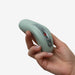 A hand holding the Fun Factory Laya 3 Lay On Humping Vibrator in sage green, an ergonomically designed electronic device with a ribbed texture. Made of body-safe silicone, it features a small control panel with buttons and a display screen.