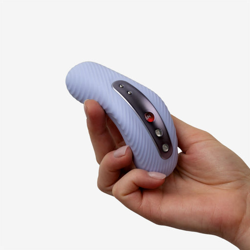 A hand holding the Fun Factory Laya 3 Lay On Humping Vibrator in Soft Violet, a small, curved, ribbed white electronic device made from body-safe silicone with a black strip on top featuring three metal buttons and a red power button. The simplistic background is a plain light gray color.
