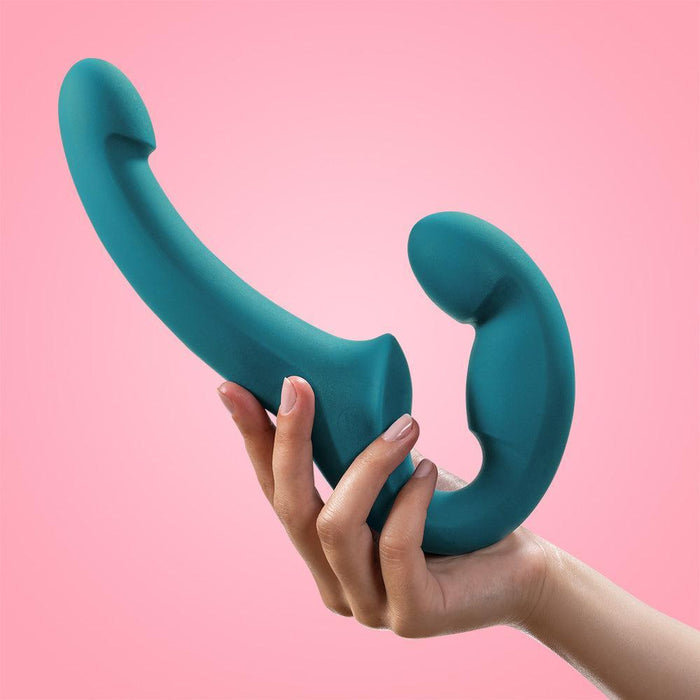 A hand is holding the Fun Factory Share Lite Silicone Double Dildo in Deep Sea Blue against a pink background. Ideal for pegging, this sex toy from Fun Factory features two distinct ends; one is slightly curved, while the other forms a pronounced hook shape.