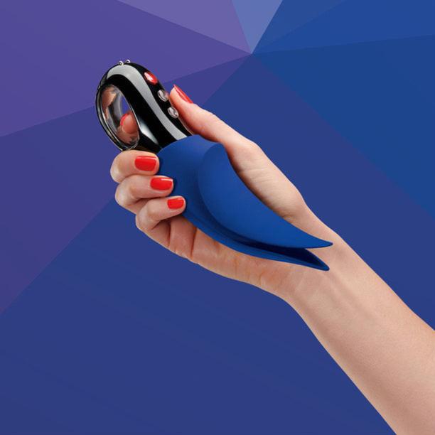 A hand with red-painted nails holds the Fun Factory Volta Rechargeable External Vibrator in Petrol Blue, showcasing its powerful motor. Its buttons suggest versatile external stimulation functions, set against a blue geometric pattern that complements its design.