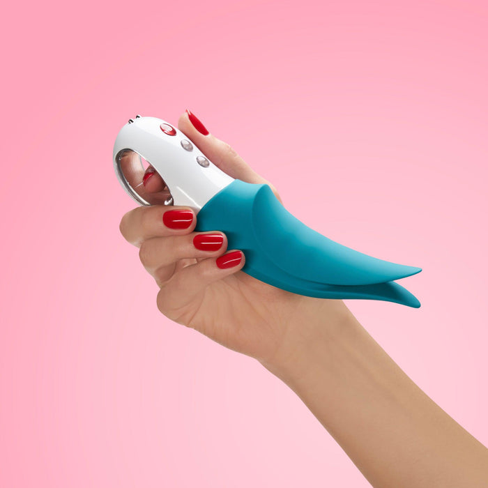 A hand with red painted nails holds a Fun Factory Volta Rechargeable External Vibrator in Petrol Blue against a pink background. The vibrator, featuring a sleek, modern design, appears to be made of metal and plastic.