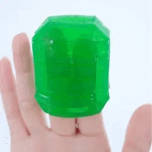 A green, transparent, gelatinous cube wobbles on a person's finger against a white background. The elasticity and texture of the Tenga Uni Emerald Textured Finger Sleeve for Stroking and Clit Massage are emphasized by its movement.