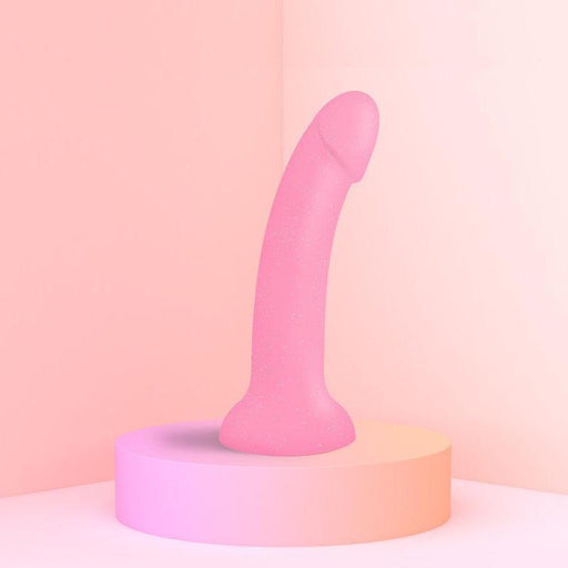 The Glitzy Pink Sparkle 7 inch Silicone Dildo by Lovely Planet, featuring a strong suction cup, is displayed upright on a circular platform against a soft gradient of pink and peach hues.