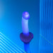 The Glow Blue LED 7 Inch Light Up Silicone Dildo stands upright on a reflective blue surface, surrounded by translucent blue panels and dramatic lighting that showcases its vibrant glow and strap-on compatible base.