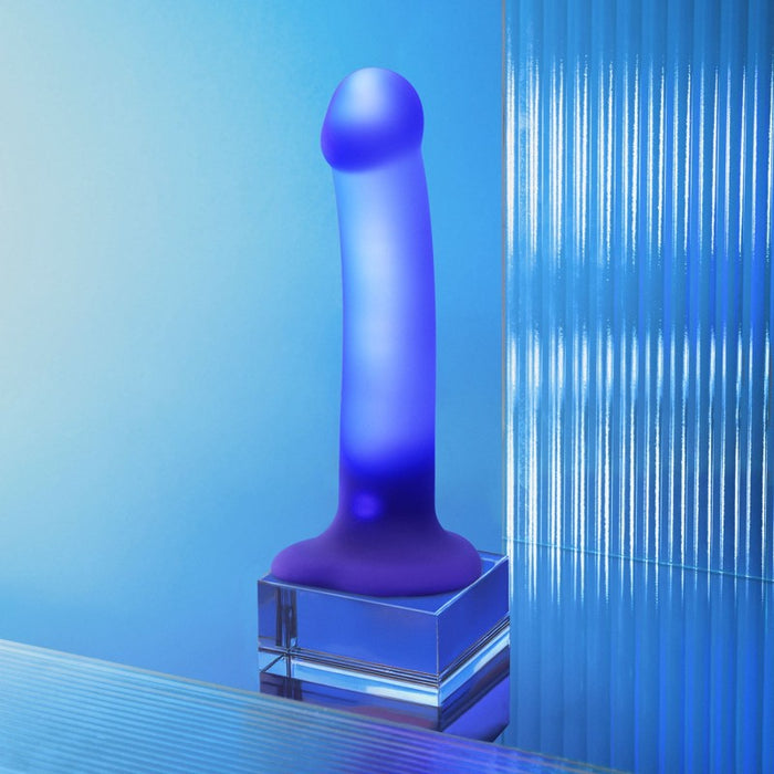 The Glow Blue LED 7 Inch Light Up Silicone Dildo stands upright on a clear square base against a modern, blue-toned background with vertical textured glass. This strap-on compatible toy combines striking style and versatile features.