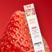 A close-up of a ripe strawberry with a box of Hot Kiss Arousal Lip Gloss - Strawberry Flavored leaning against it, set on a vibrant red background. The packaging promotes sugared lips and its sugar-free formula in English and French.