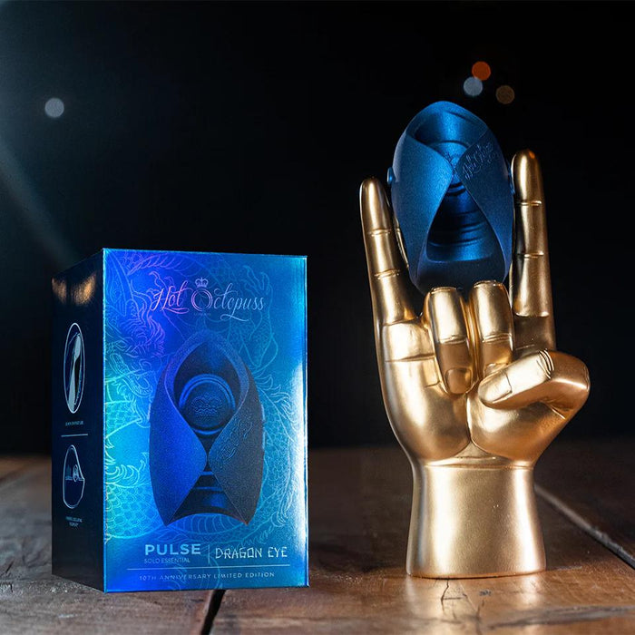 A blue box labeled "Hot Octopuss Pulse Solo Dragon Eye Oscillating Penis Masturbator" stands beside a gold “rock on” hand sculpture with a dark blue Guybrator balanced on its fingers, displayed on a wooden surface.