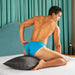 A man in bright blue underwear kneels on a bed, posing with one knee on the Liberator Humphrey Pillow-Style Sex Toy Mount. He looks back over his shoulder, with teal curtains and a headboard visible in the background.