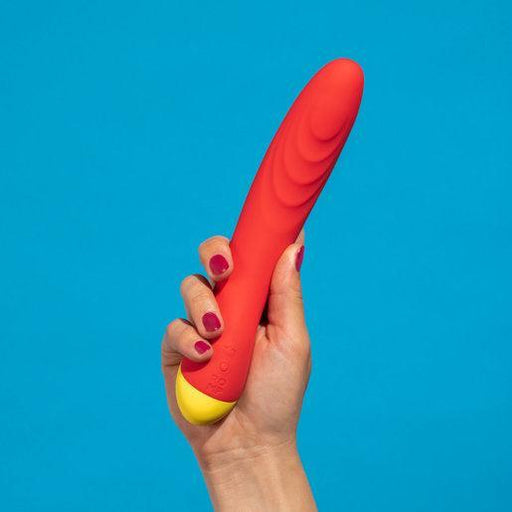 A hand with red-painted nails holds a sleek Hype Beginner's Silicone G-Spot Vibrator by WOW in vibrant red and yellow against a striking blue background.