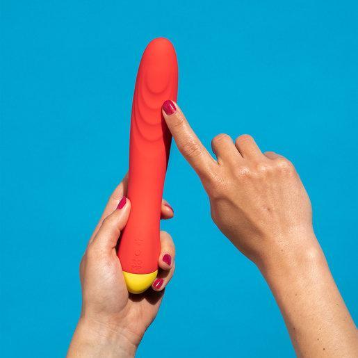 A person with red-painted nails is holding the WOW Hype Beginner's Silicone G-Spot Vibrator, which features a striking red and yellow color scheme, against a bright blue background. The ergonomic design is emphasized as the person points at the device with their other hand.