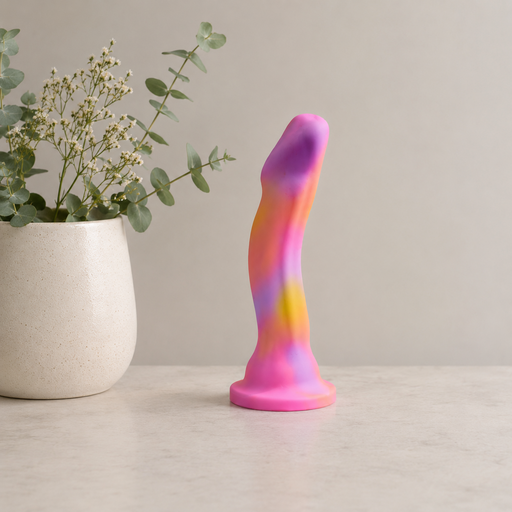 The Sun's Out 7.5 Inch Pink Ombre Silicone G Spot Dildo, featuring pink, yellow, and purple hues, stands upright on a light surface beside a white vase filled with green eucalyptus sprigs and small white flowers.