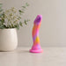 The Sun's Out 7.5 Inch Pink Ombre Silicone G Spot Dildo, featuring pink, yellow, and purple hues, stands upright on a light surface beside a white vase filled with green eucalyptus sprigs and small white flowers.