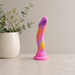 The Sun's Out 7.5 Inch Pink Ombre Silicone G Spot Dildo, featuring pink, yellow, and purple hues, stands upright on a light surface beside a white vase filled with green eucalyptus sprigs and small white flowers.