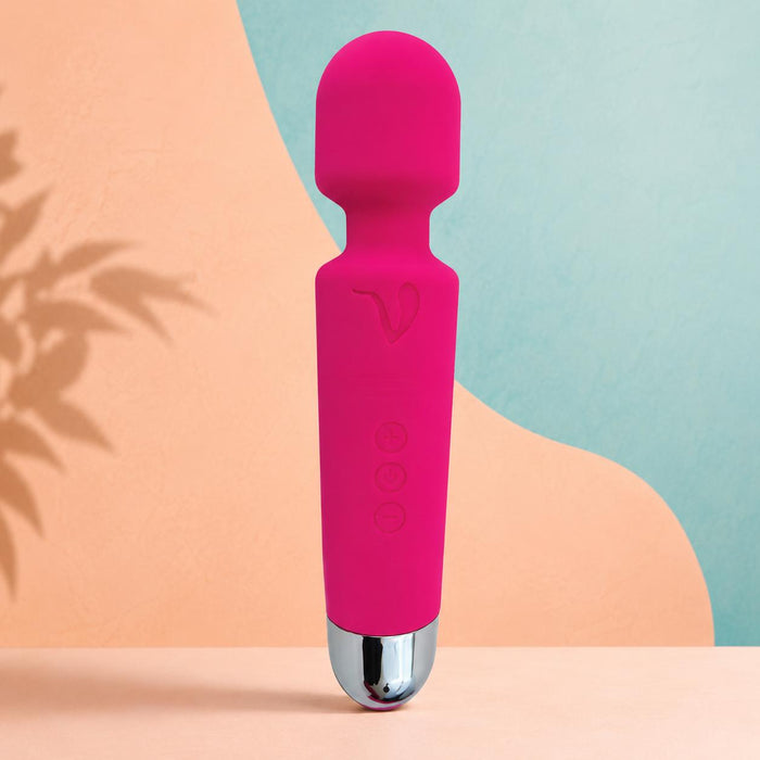 The Mini Halo Extra Powerful Wand Vibrator in bright pink, featuring a rounded top and silver base, stands upright on a peach surface with a pastel blue and peach abstract background.