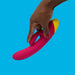 A hand holding the Jazz Beginner's Silicone Rabbit Vibrator by WOW, with pink and yellow silicone, against a blue background. The person with brown skin and red painted nails showcases this beginner-friendly vibrator designed for G-spot stimulation.