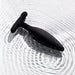 The Je Joue Egon Powerful Remote Control Vibrating Butt Plug, crafted from sleek black silicone with a handle, sits on a white surface as gentle circular ripples form in the water around it.