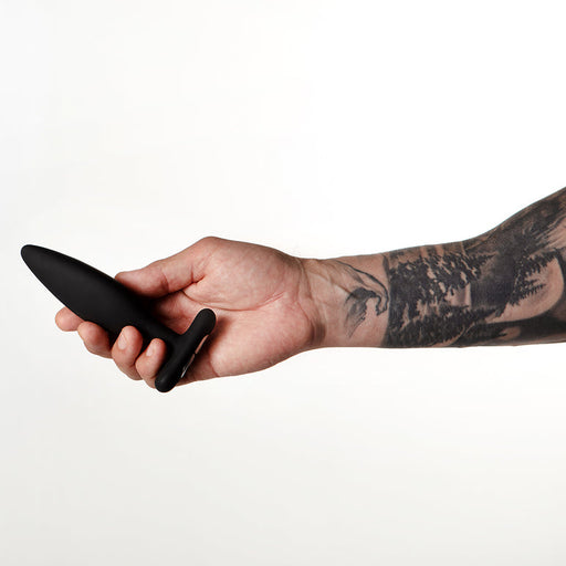 A tattooed arm holds the Je Joue Egon Powerful Remote Control Vibrating Butt Plug, made of black silicone, against a plain white background.