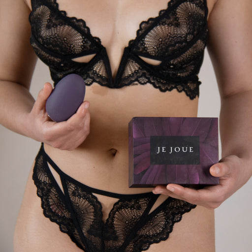 A woman dressed in black lace lingerie holds a Je Joue Mimi Clitoral and External Vibrator - Purple, an egg-shaped item known for its exceptional clitoral stimulation, in her right hand and a purple box labeled "Je Joue" in her left hand. The background is neutral and uncluttered.