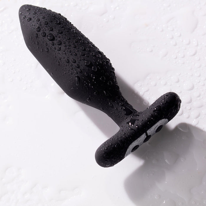 The Je Joue Onyx Large Powerful Remote Control Vibrating Butt Plug, in black silicone and built for deep rumbly vibrations, rests on a white surface glistening with water droplets.