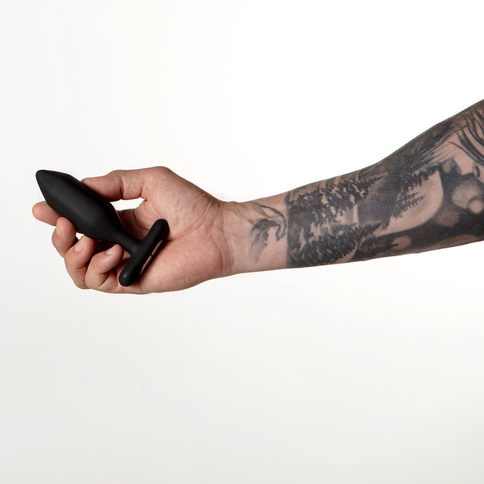 A tattooed arm holds the Je Joue Onyx Large Powerful Remote Control Vibrating Butt Plug, black in color, against a plain white background.