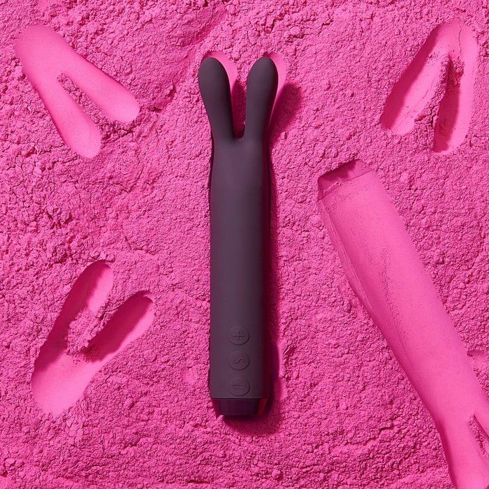 A purple, rabbit-ear-shaped Je Joue Powerful Bullet with Flexible Rabbit Ears lies on a textured pink surface. It is surrounded by bunny footprint imprints in the pink material. The vibrator has control buttons at the base, designed to deliver rumbly vibrations for your pleasure.