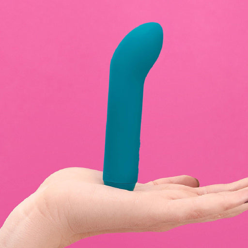 The Je Joue Powerful Internal or External Bullet in teal, a premium silicone vibrator with a sleek and smooth waterproof design, is balanced upright on a person's open palm against a solid pink background.