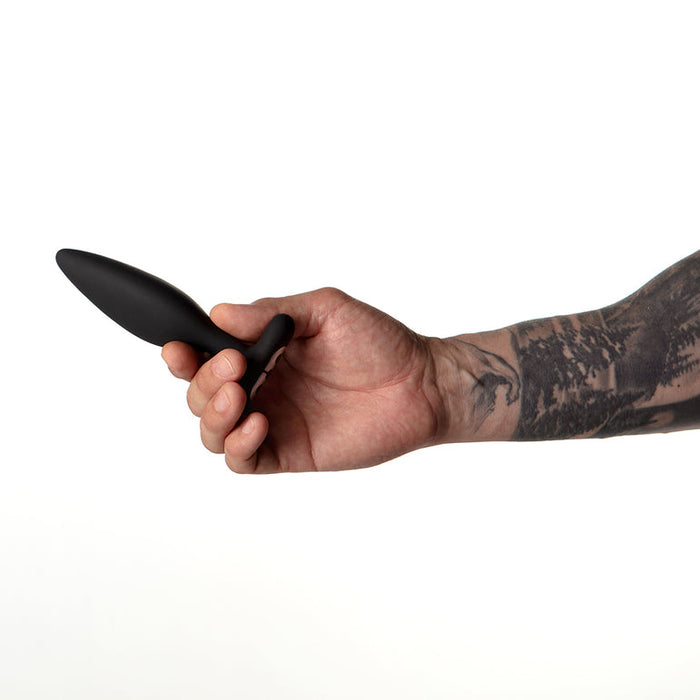 A tattooed hand holds the Je Joue Vesta Petite Powerful Remote Control Vibrating Butt Plug, made from body-safe silicone, against a plain white background.